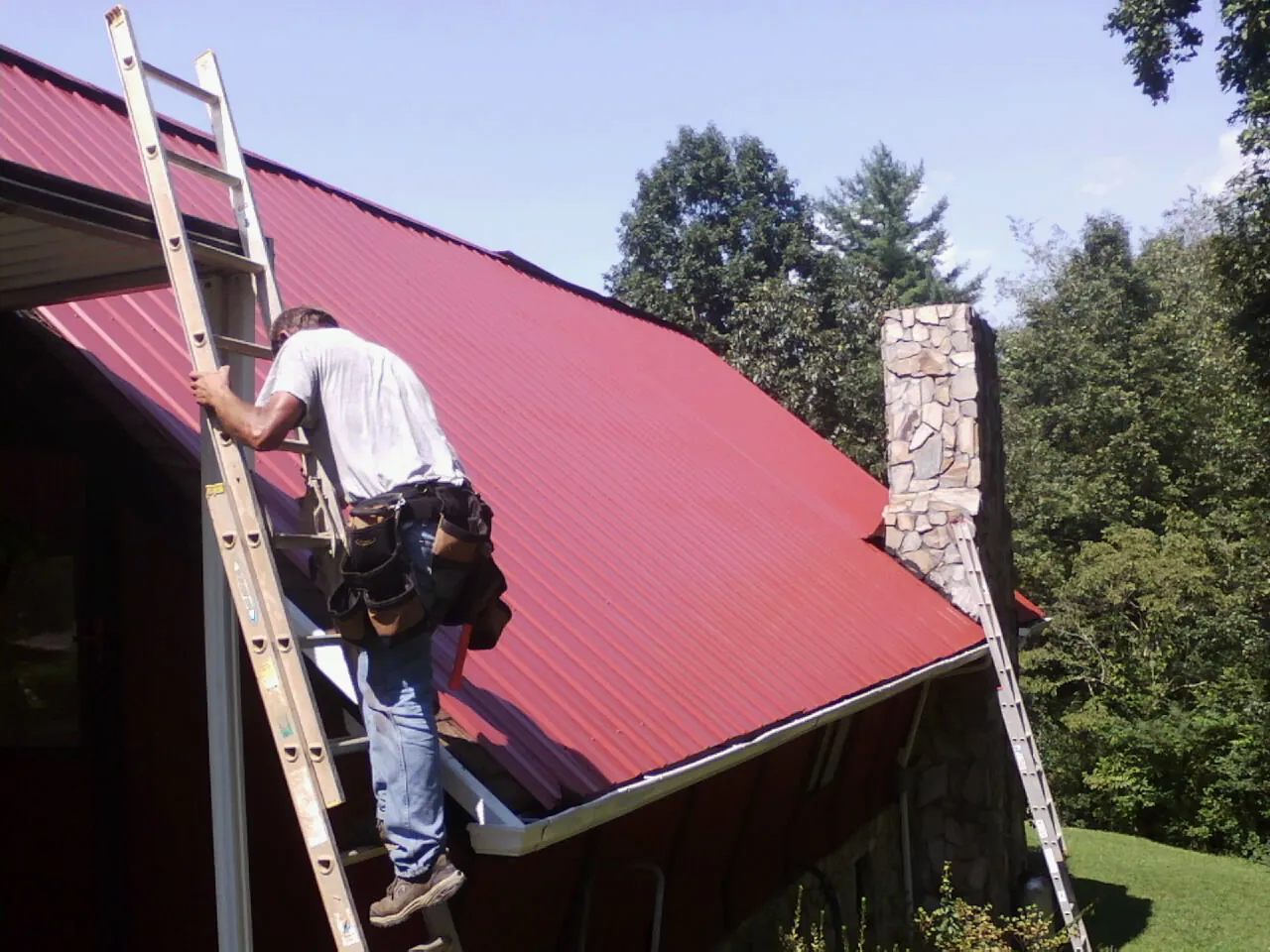 Roofer on a ladder beside a red metal roof and stone chimney — Soffit & Fascia Repair by Roofing Brothers in Williston Park